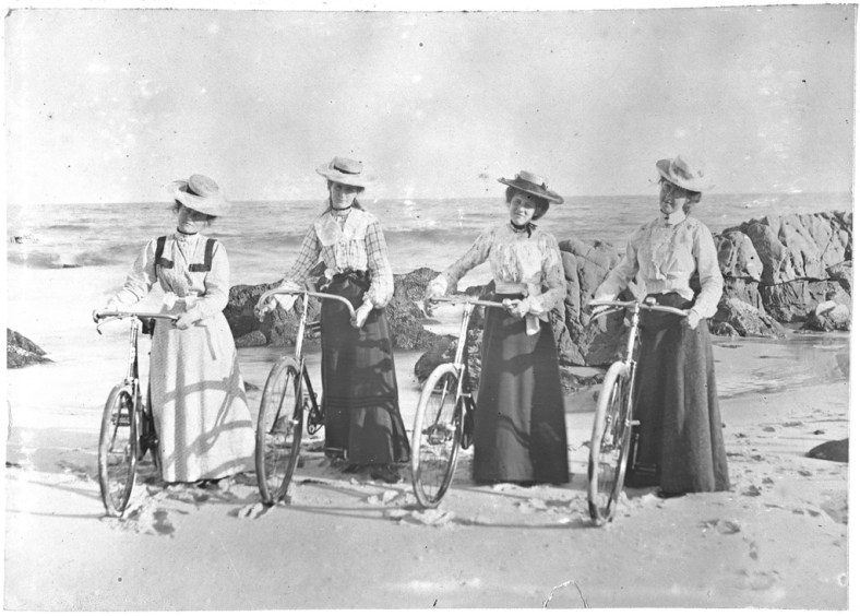 Four women on a beach with bicycles.